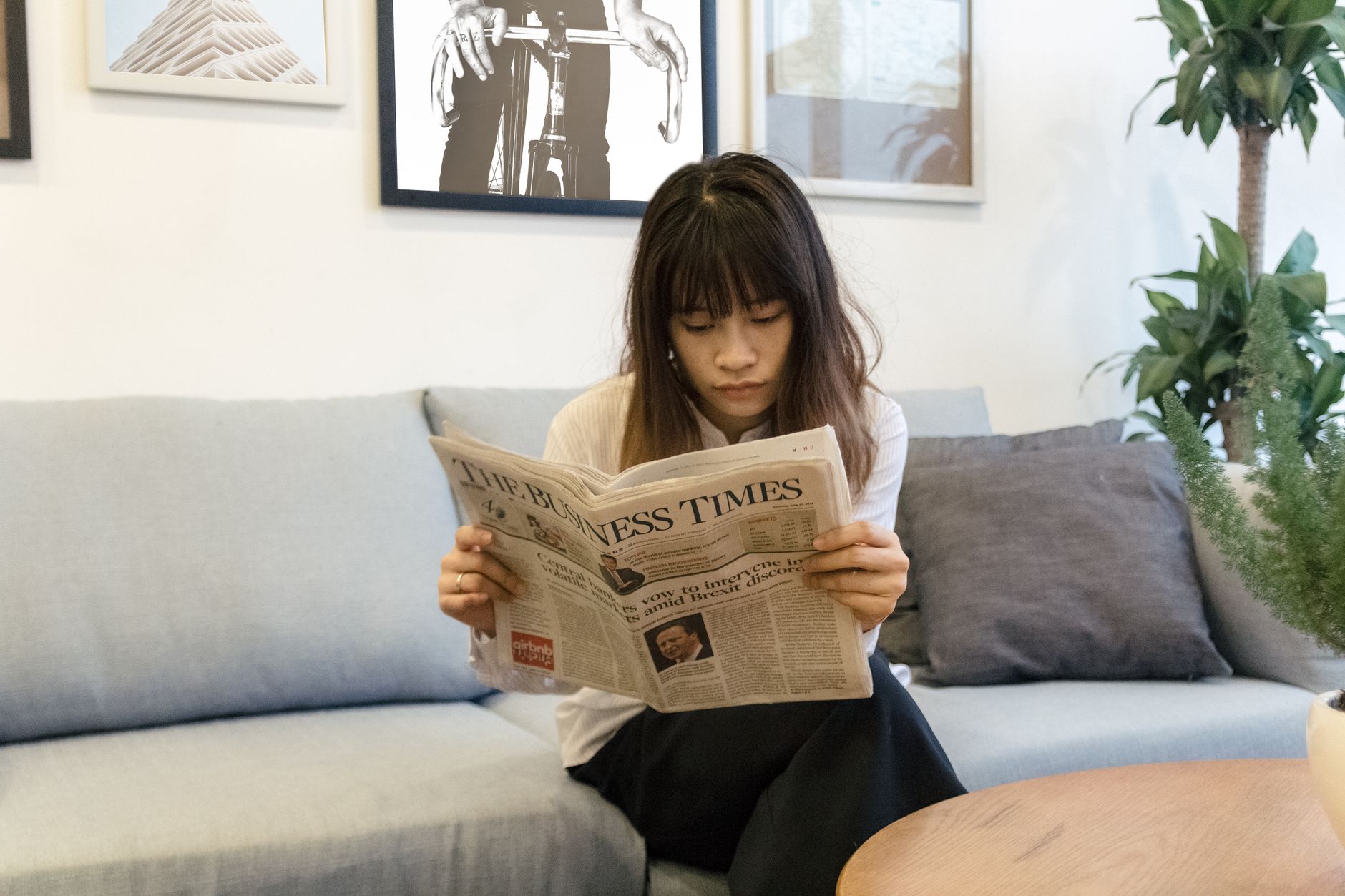 A young woman attentively reads a newspaper while sitting on a modern sofa indoors. — test123123